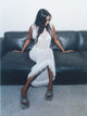 A model with both arms pushing down on a dark couch looking off in one direction wearing the 08 Fringe Dress in white