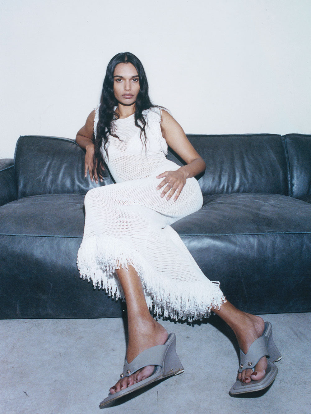 A model relaxing on a dark couch wearing the 08 Fringe Dress in white