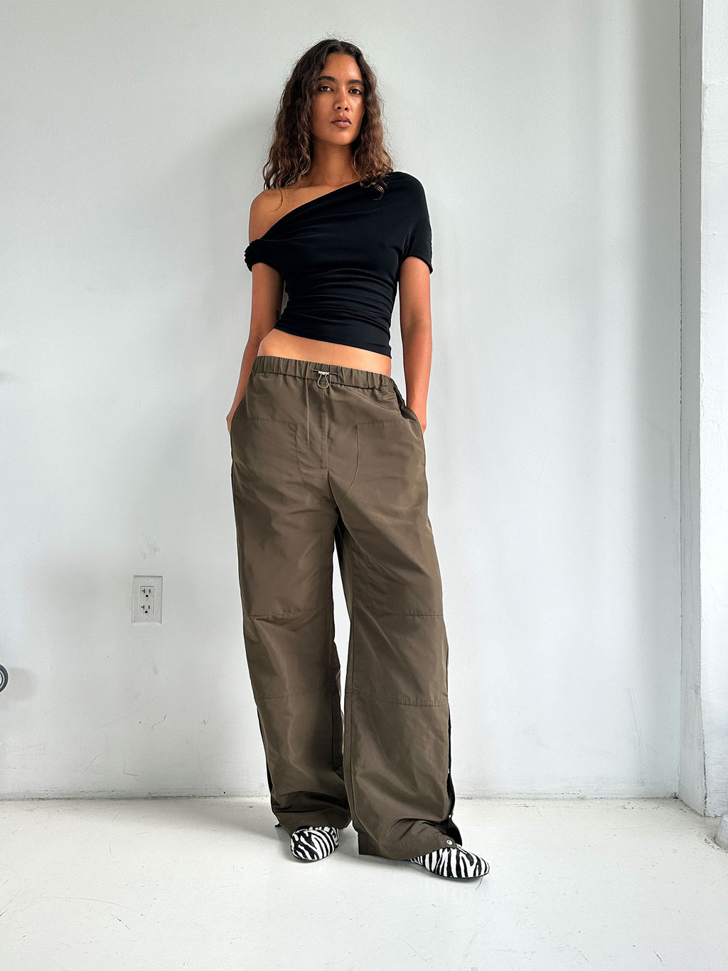 A model standing facing forwards wearing the Black Elemental Ida Top with her hands in the pockets of the Elemental Parachute Pants in Seaweed