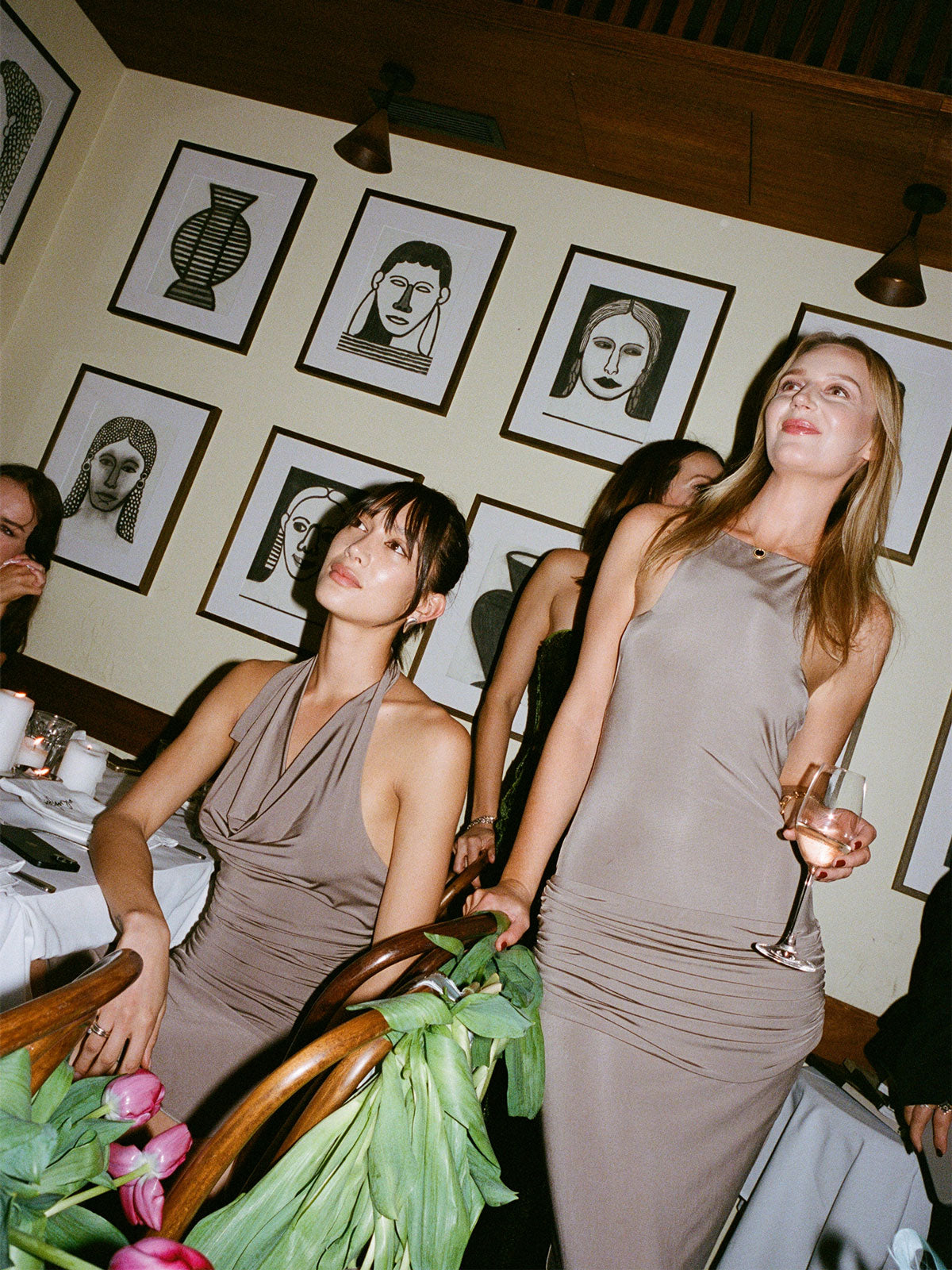 Two female figures at an event with one sitting down and the other standing holding her drink and one is wearing the Concrete 09 Danae Dress