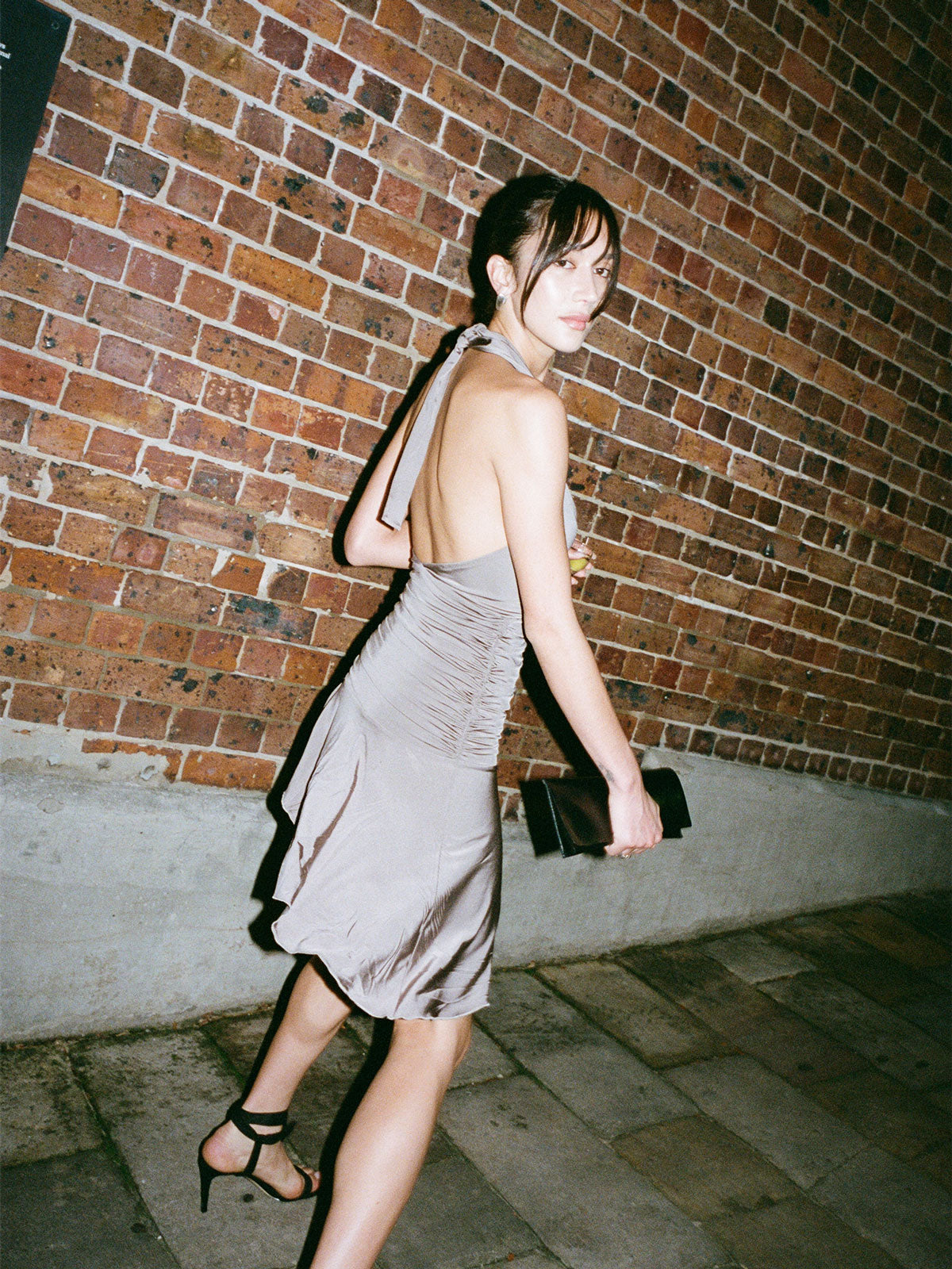 A model walking near a brick wall and cobbled street looking off to the side wearing the Concrete 09 Halter Mini and holding her purse